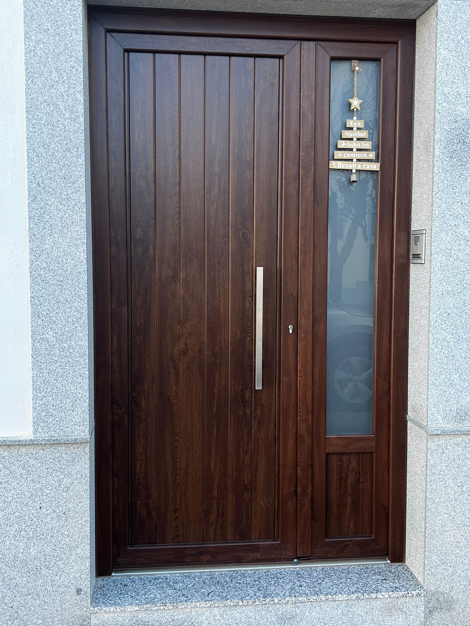 Puerta principal de madera con un largo tirador plateado y un panel de vidrio esmerilado, empotrada en una pared exterior de piedra