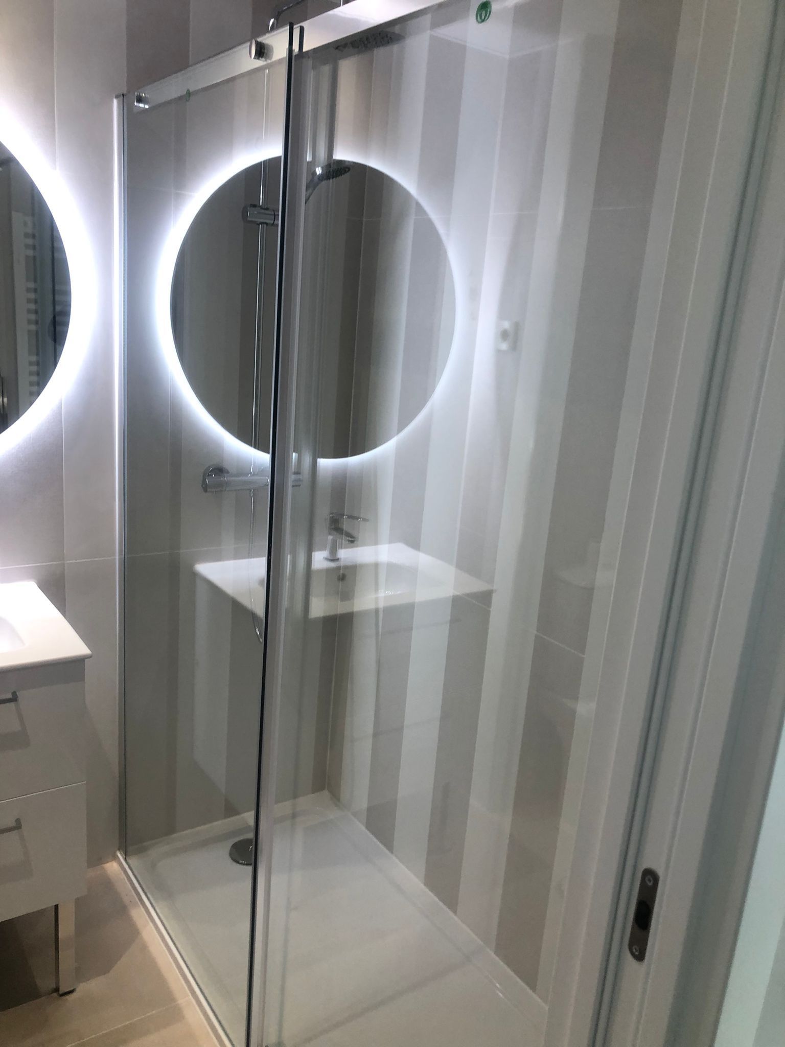 Baño moderno con ducha y mampara de cristal, espejo redondo retroiluminado y encimera de lavabo blanca.