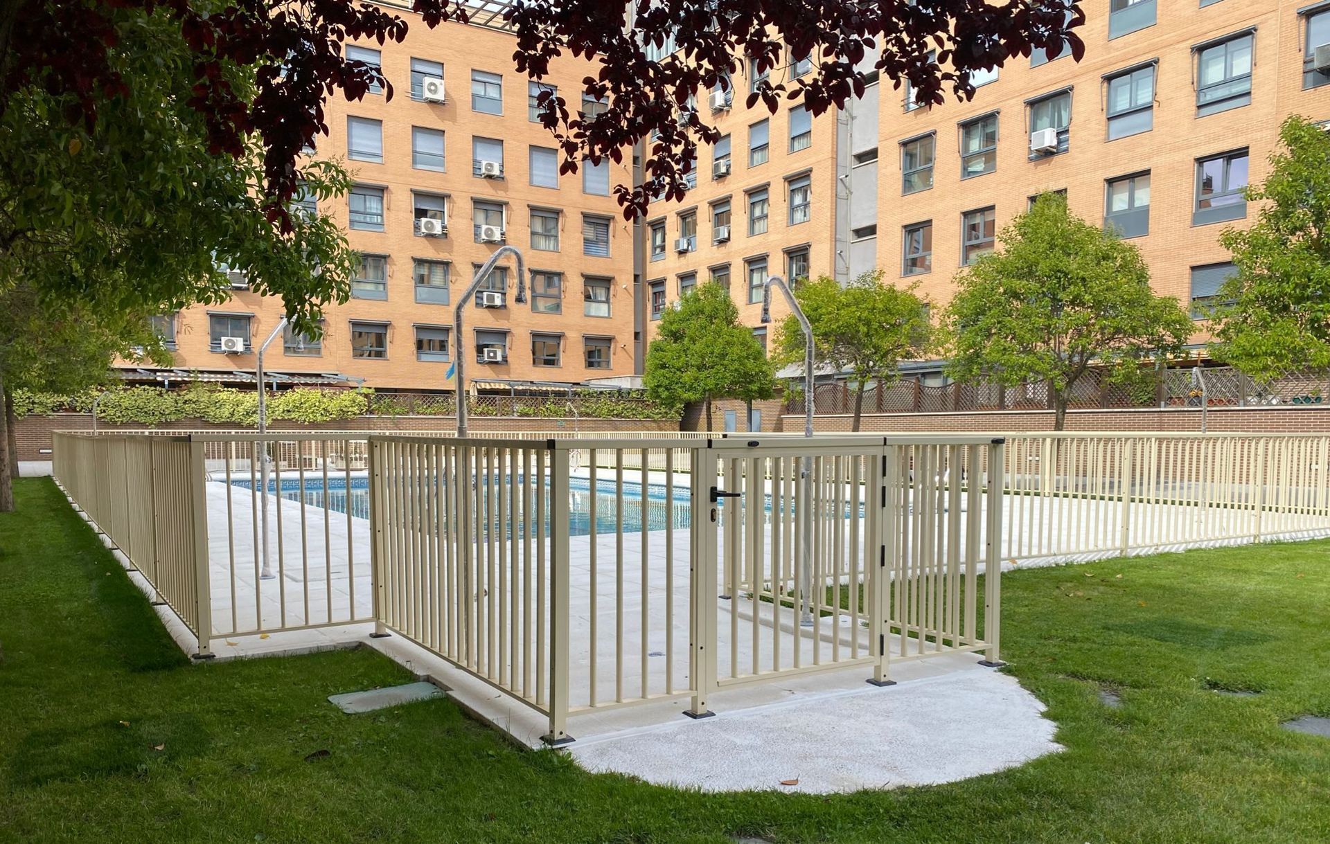 Piscina exterior rodeada de una valla blanca, junto a un edificio de apartamentos color beige y un patio con césped.