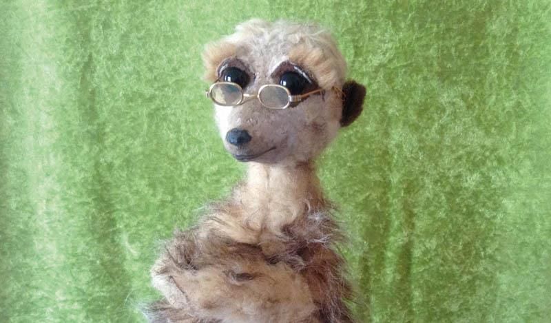 Ein ausgestopftes Erdmännchen mit Brille steht auf einem grünen Tuch.