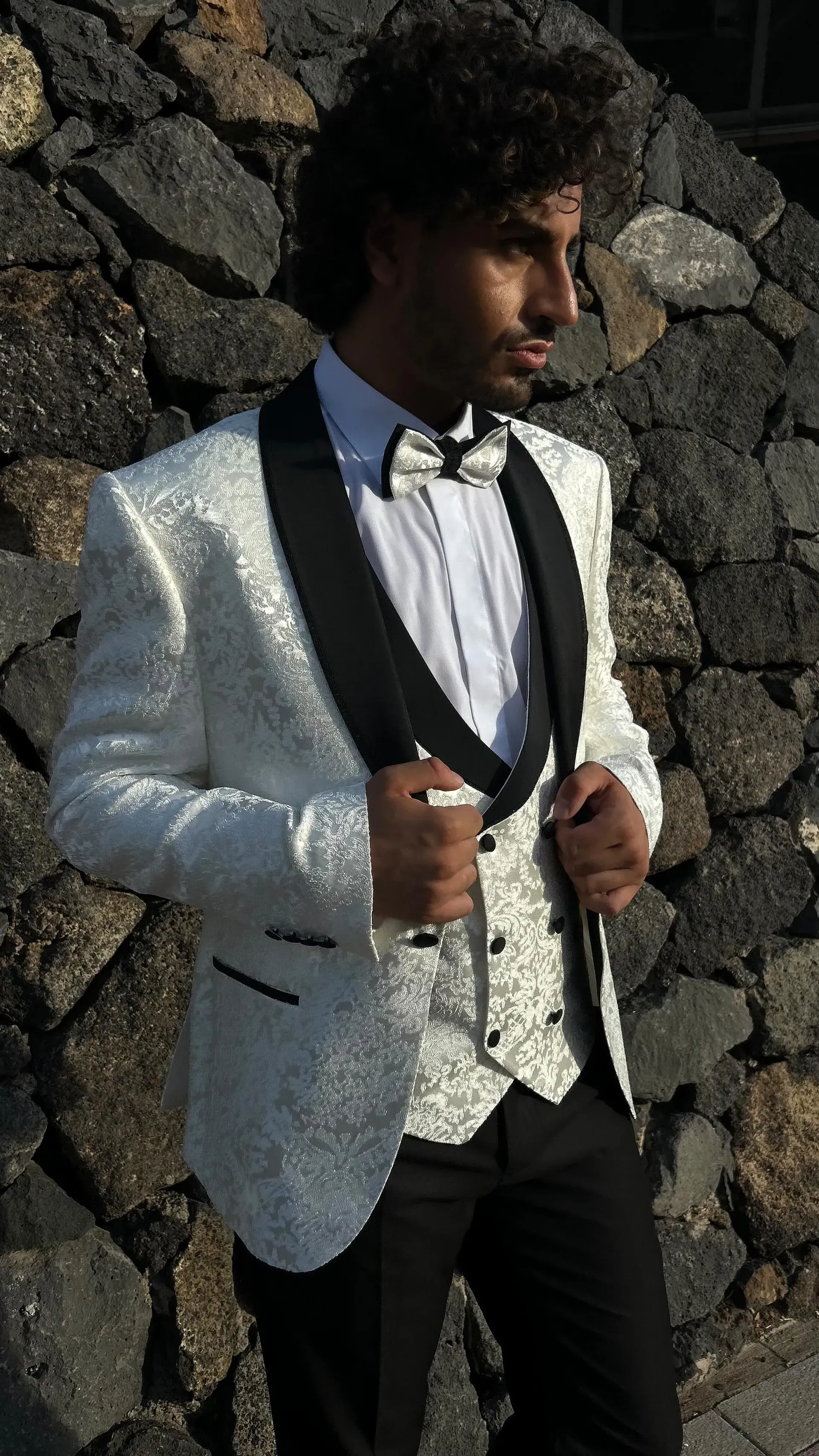 Un hombre con un esmoquin blanco y pantalones negros está parado frente a una pared de roca.