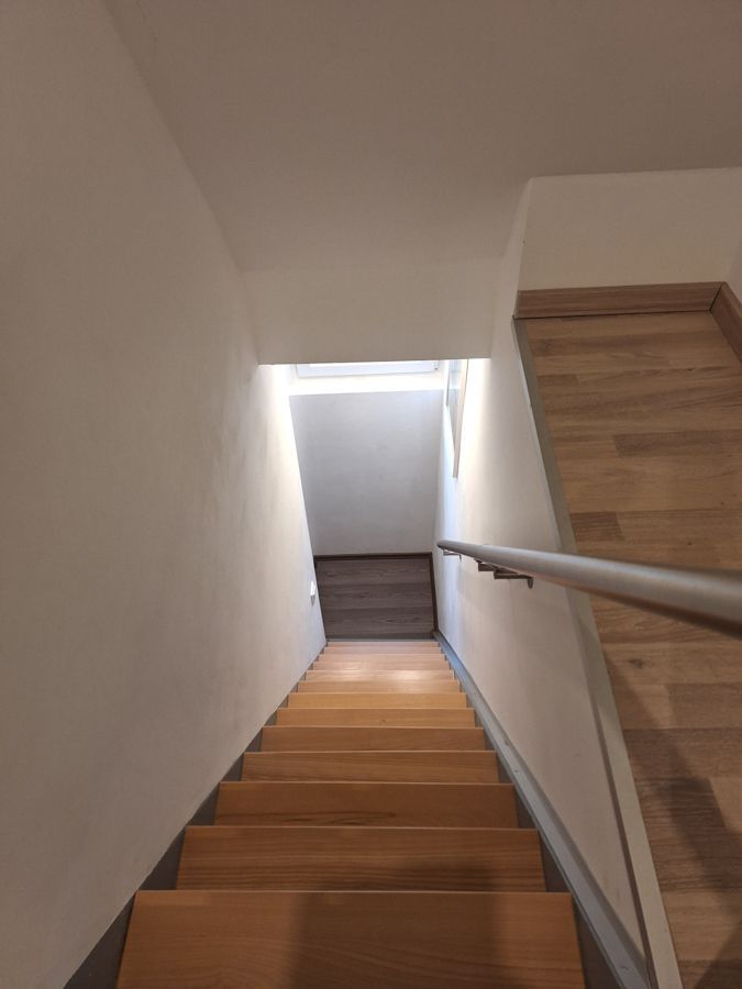 Nach unten führende Holztreppe; weiße Wände, Metallhandlauf und Licht am Ende.