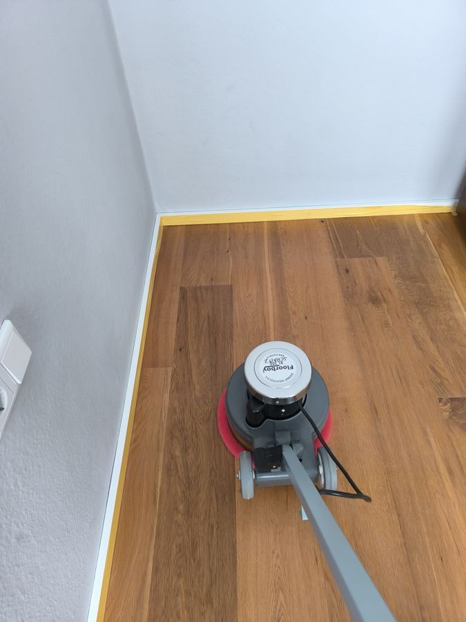Bodenpoliermaschine reinigt einen Holzboden neben einer grauen Wand mit einem Lichtschalter.