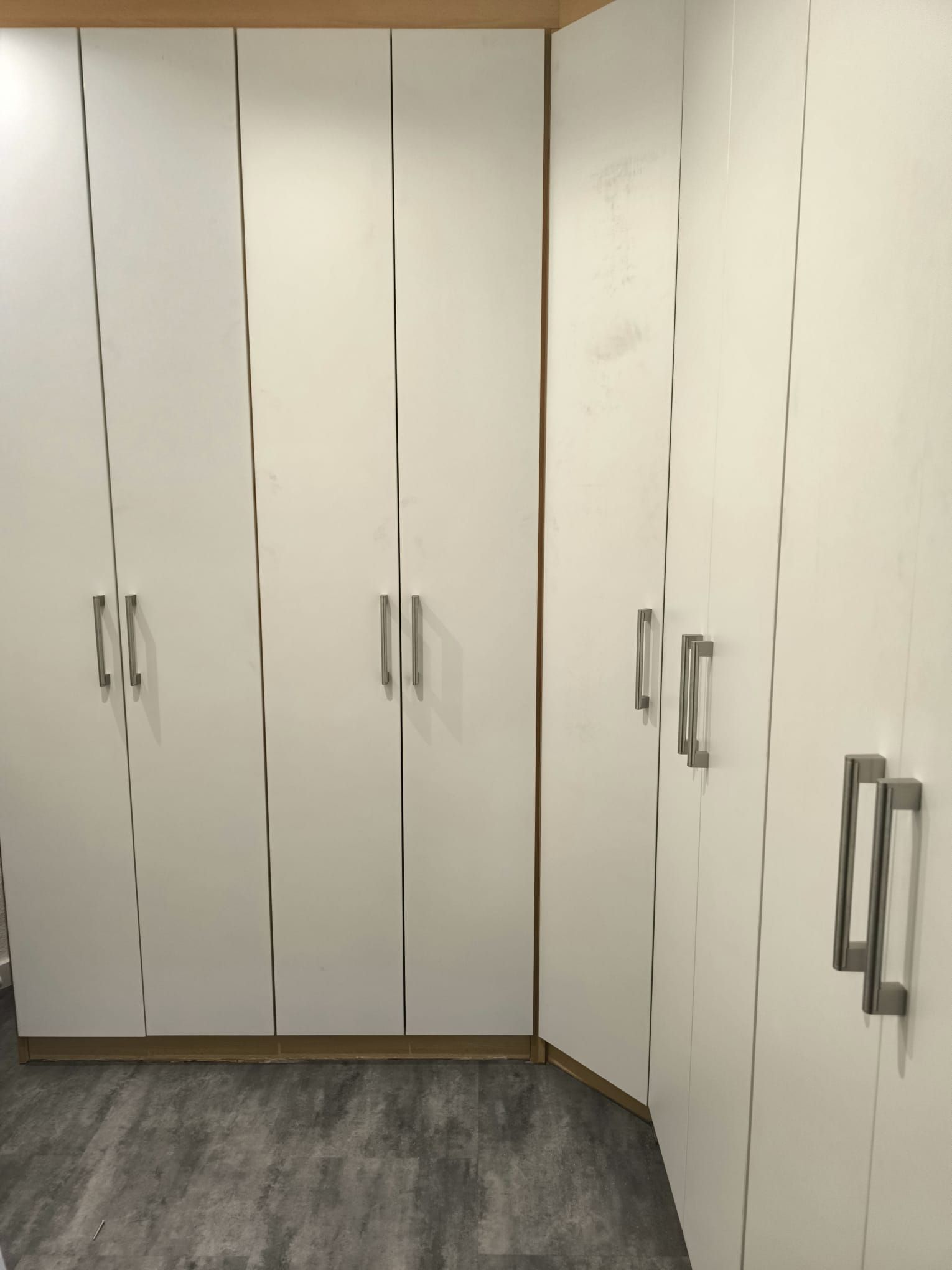 Weiße Schranktüren mit silbernen Griffen in einem Eckzimmer mit grauem Bodenbelag.