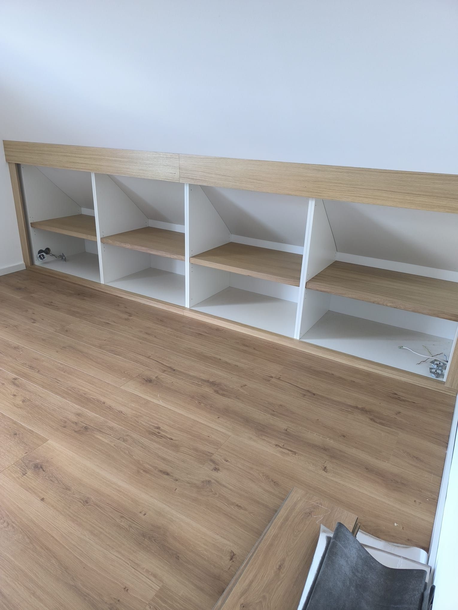 Eingebautes weißes Regal mit Holzböden und -platte vor einer weißen Schrägwand über einem Holzboden.