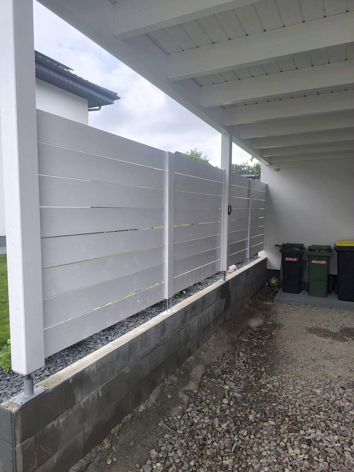 Ein weißer Zaun umgibt eine Garage mit Mülltonnen.