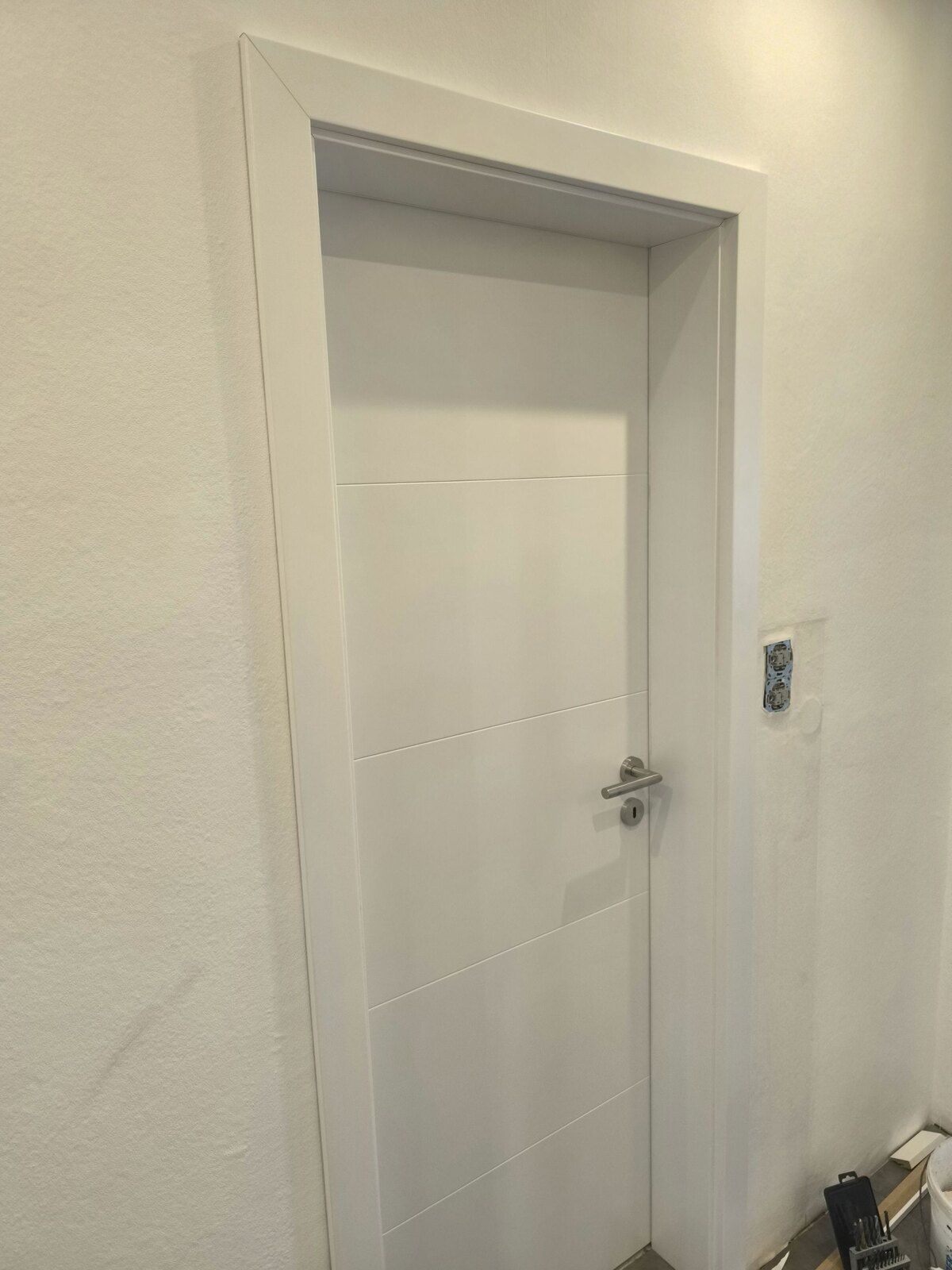 In einem Flur steht neben einer weißen Wand eine weiße Tür.