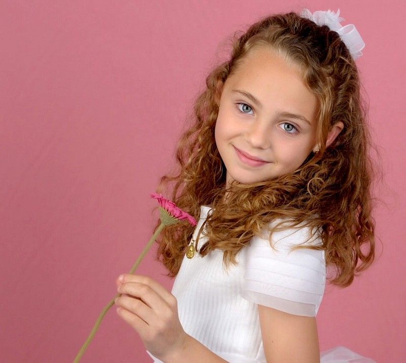 Una chica con cabello rizado, sosteniendo una flor rosa, sonríe a la cámara sobre un fondo rosa.
