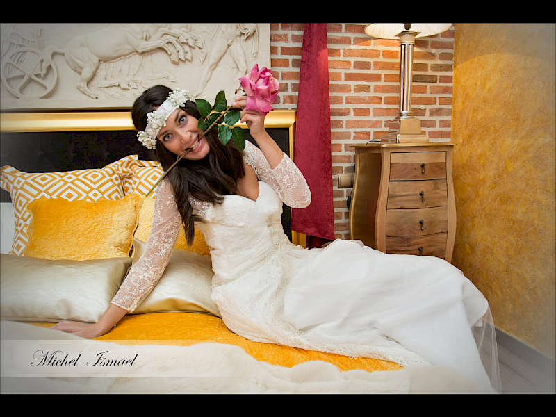Una mujer con vestido de novia, sosteniendo una rosa rosa, sonríe. Está sentada en una cama con detalles amarillos.