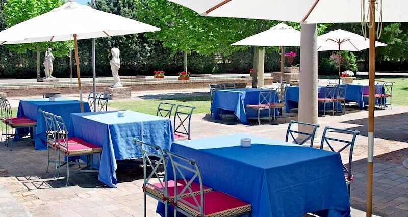 Mesas de comedor al aire libre con manteles azules, sillas rosas y sombrillas blancas. Jardín.