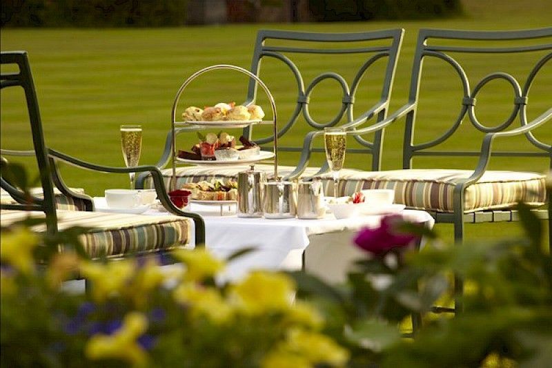 Juego de té al aire libre con pasteles, champán y sillas de metal en un césped, soleado.
