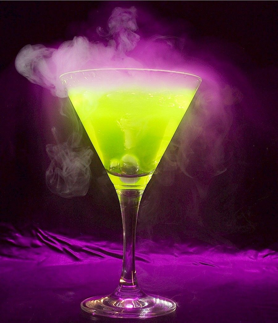 Cóctel verde en copa de martini, con humo elevándose sobre un fondo morado.