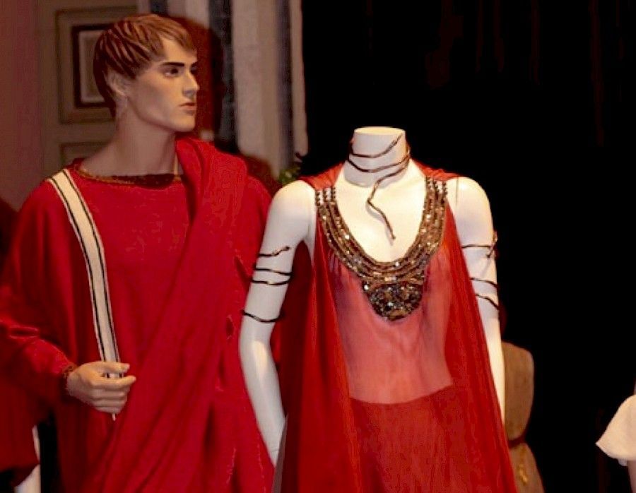 Dos maniquíes vestidos con ropas rojas de estilo romano: uno con túnica y capa, el otro con vestido transparente y joyas.