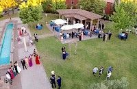 Recepción de boda al aire libre, invitados reunidos cerca de mesas bajo sombrillas, piscina y edificio al fondo.