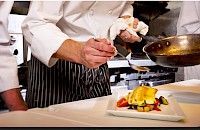 Chef preparando un plato con salsa en una cocina profesional, usando una cuchara y sosteniendo una toalla.