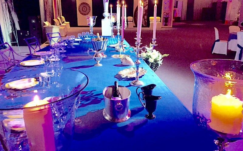 Mesa larga con mantel azul, preparada para un evento, adornada con velas y cubiertos.