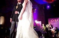 Una pareja con traje formal en el escenario de un evento. La novia lleva vestido blanco y velo; el novio.
