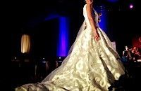 Mujer con un vestido de novia blanco con una larga cola en un escenario, fondo azul y oscuro.