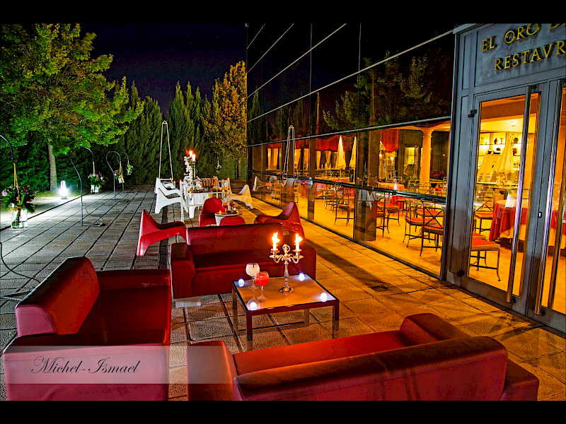 Patio exterior de noche con sofás rojos, mesas pequeñas y reflejo del restaurante.