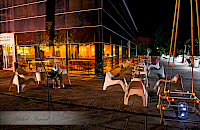 Cafetería al aire libre de noche. Sillas blancas en una plaza, con un edificio iluminado al fondo.