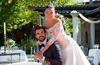Novia y novio sonriendo, abrazándose en una recepción de boda al aire libre; ella con un vestido blanco, él con un traje.