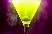 Cóctel verde en copa de martini sobre fondo morado, con un efecto blanco brumoso.