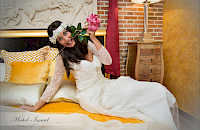 Una mujer con vestido blanco se reclina en la cama, sosteniendo una rosa rosa, sonriendo.