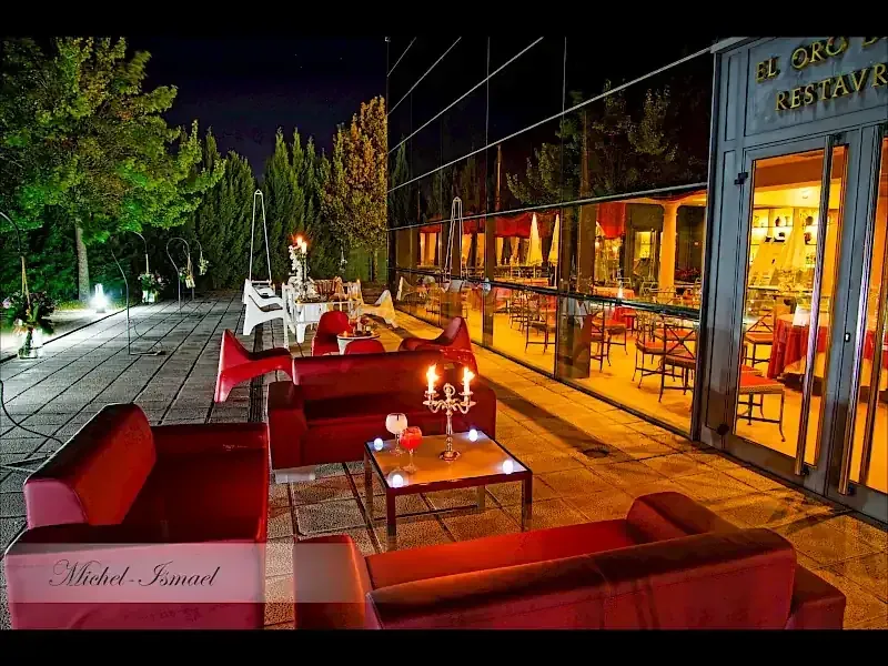 Patio exterior de noche con sofás rojos, mesas pequeñas y reflejo del restaurante.