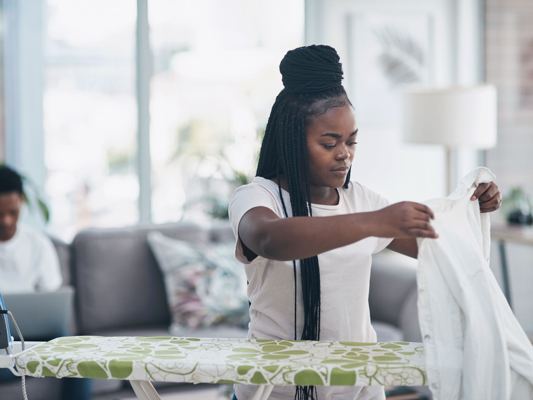 Femme pliant du linge blanc sur une planche à repasser dans un salon lumineux.