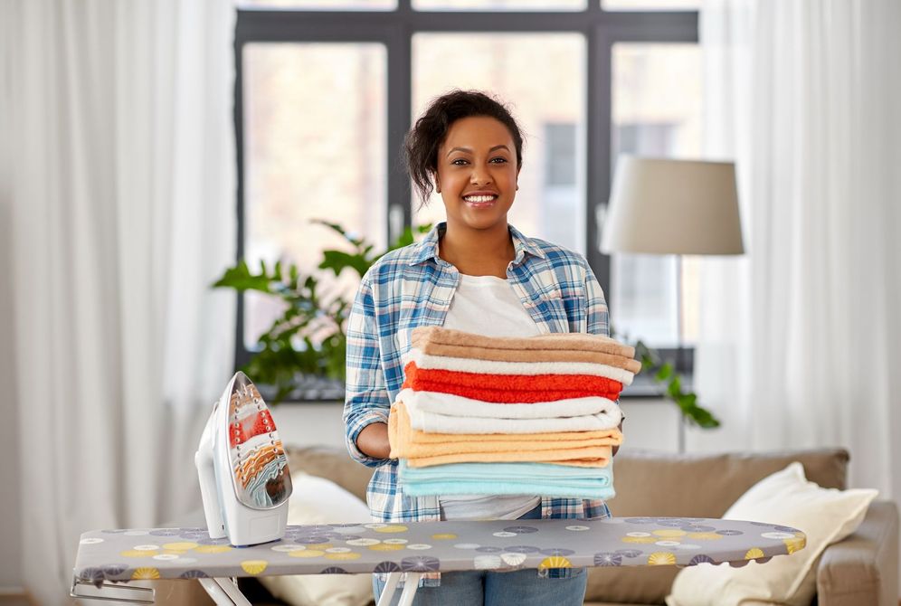 Une personne sourit à côté d'une pile de linge plié, un fer à repasser posé sur une table dans un salon lumineux.