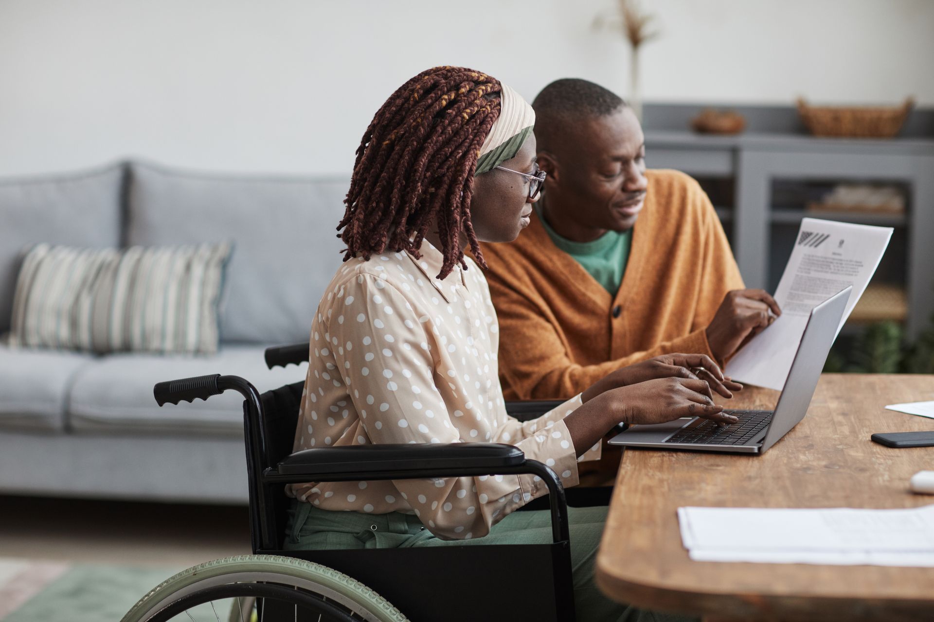 Deux personnes utilisent un ordinateur portable à une table, l'une d'elles étant en fauteuil roulant, dans un salon.