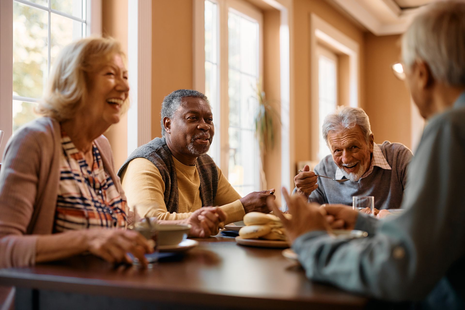 Des amis rient et partagent un repas à une table dans une pièce lumineuse.