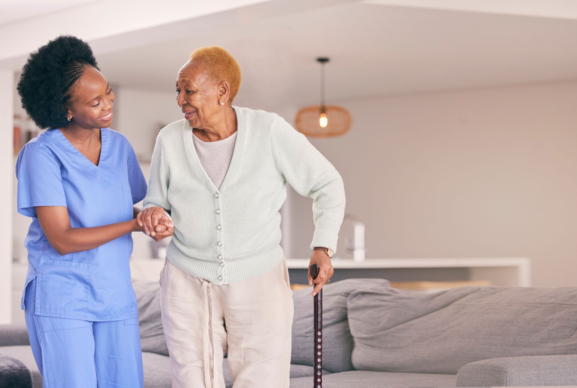 Une aide-soignante accompagne une femme âgée qui marche avec une canne dans un salon lumineux.