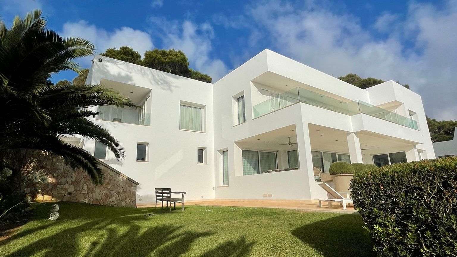 Casa moderna blanca sobre una gespa verda, amb un cel blau i palmeres.