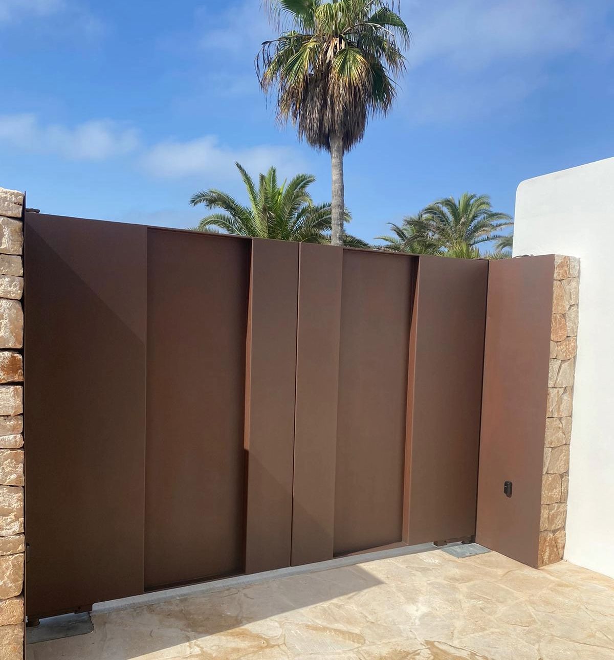 Porta metàl·lica marró amb panells verticals, flanquejada per pilars de pedra, contra un cel blau amb palmeres.