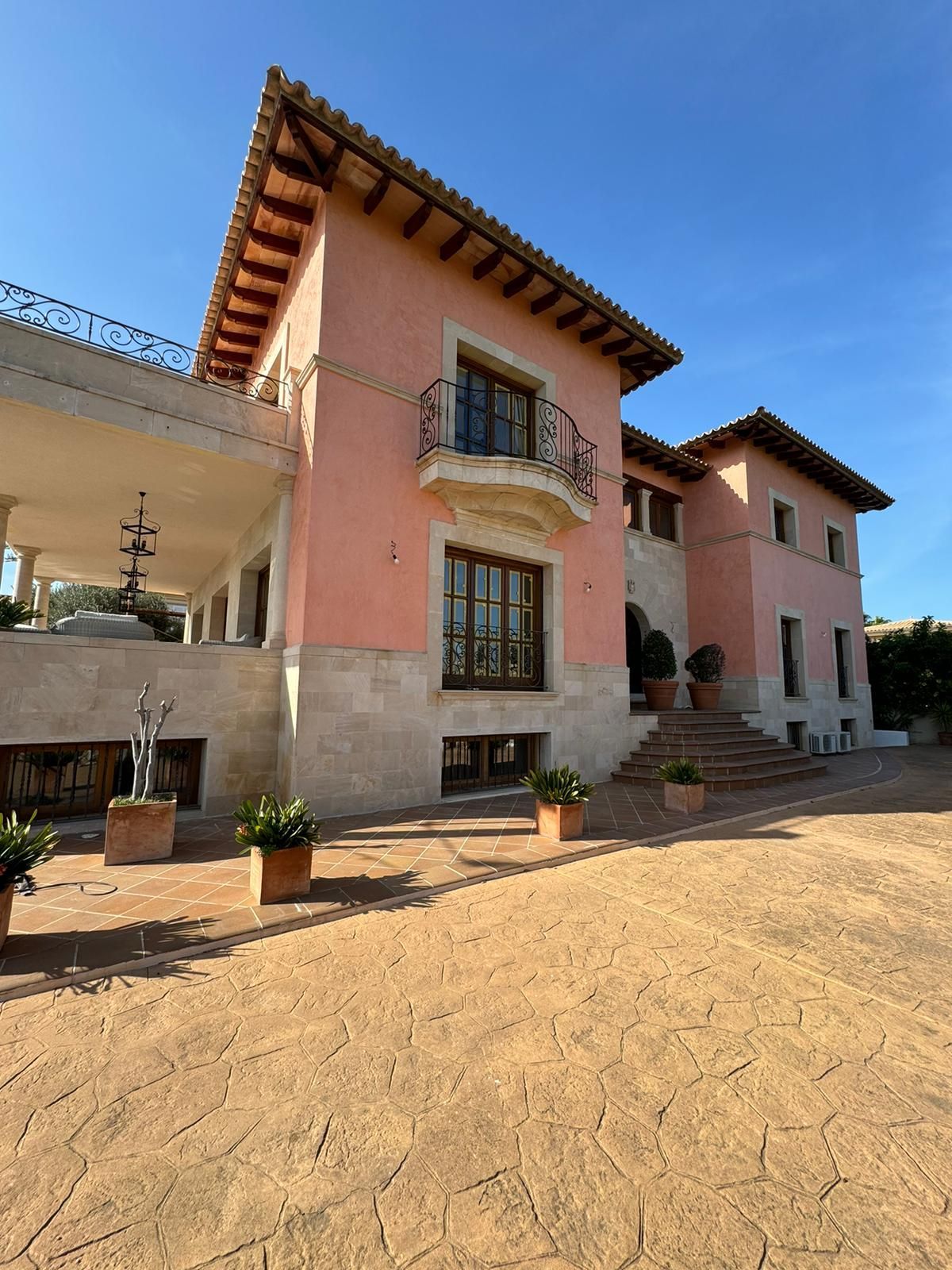 Mansió d'estuc rosa amb base de pedra, teulada de terracota i plantes en test en un entorn assolellat a l'aire lliure.