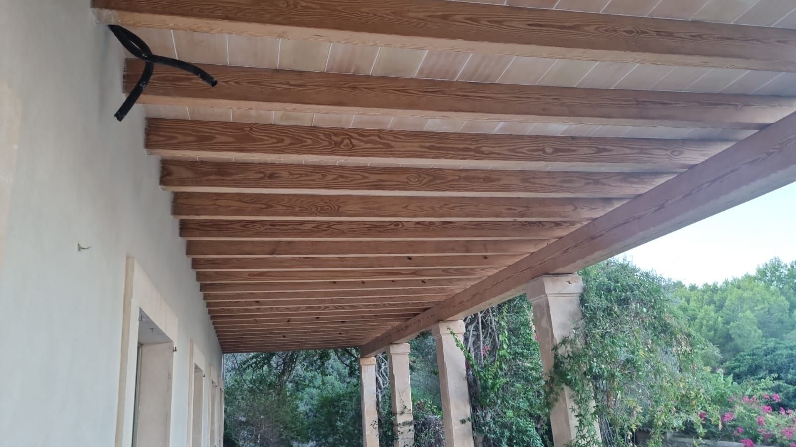 Pèrgola de fusta sobre un porxo amb pilars de pedra. Fullatge verd al fons.