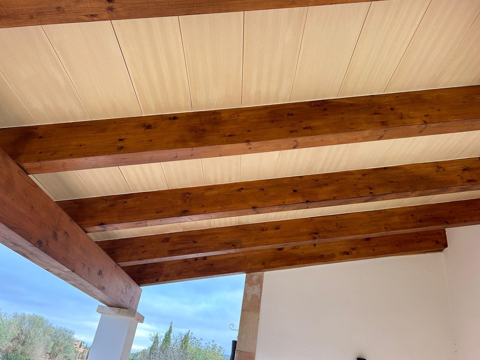 Bigues de fusta que sostenen un sostre texturat de color clar. Vista exterior, cel parcialment ennuvolat visible.