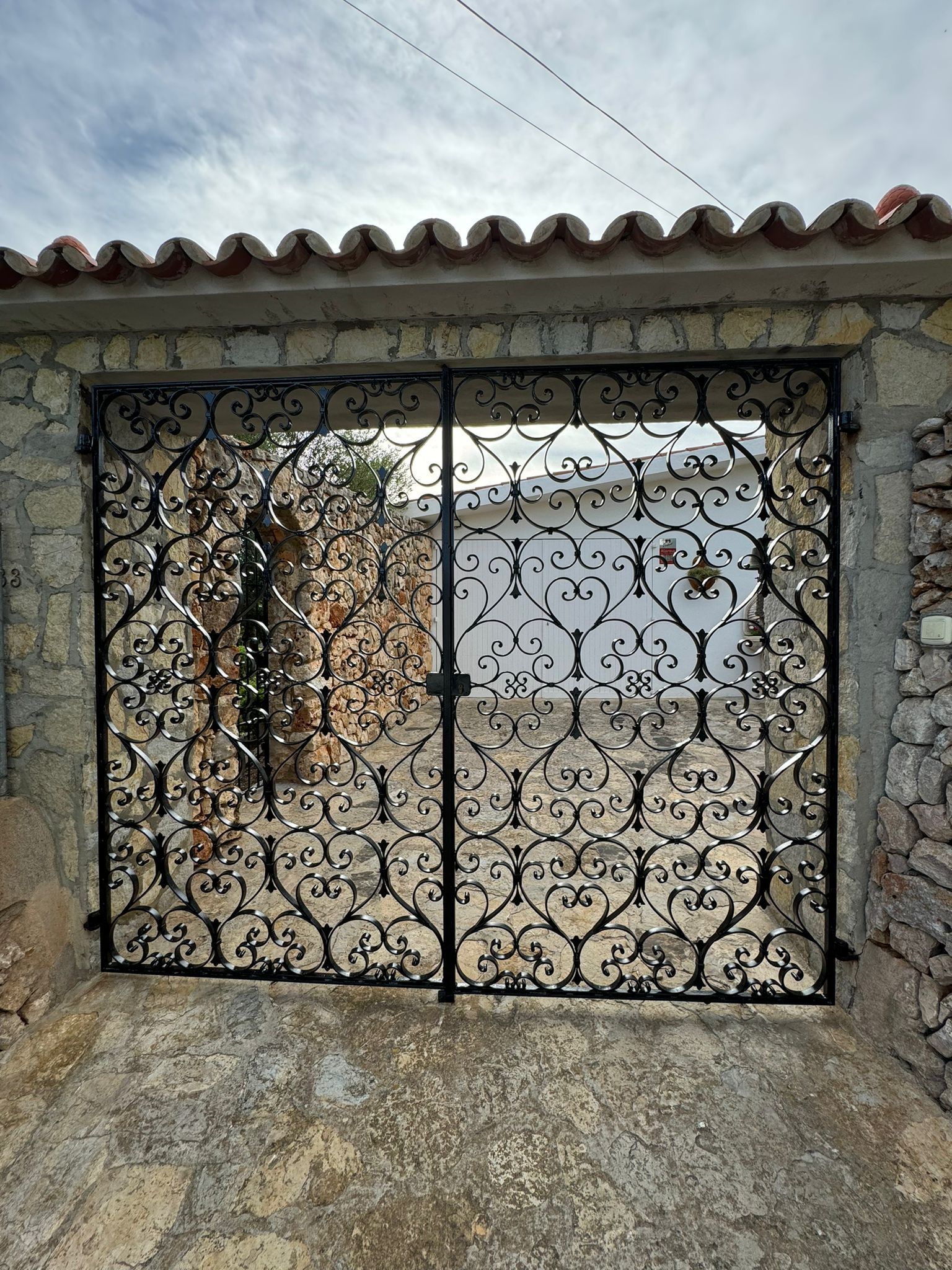 Porta de ferro forjat negre en arc de pedra, disseny floral ornamentat, tancada. Terra de formigó, cel ennuvolat.