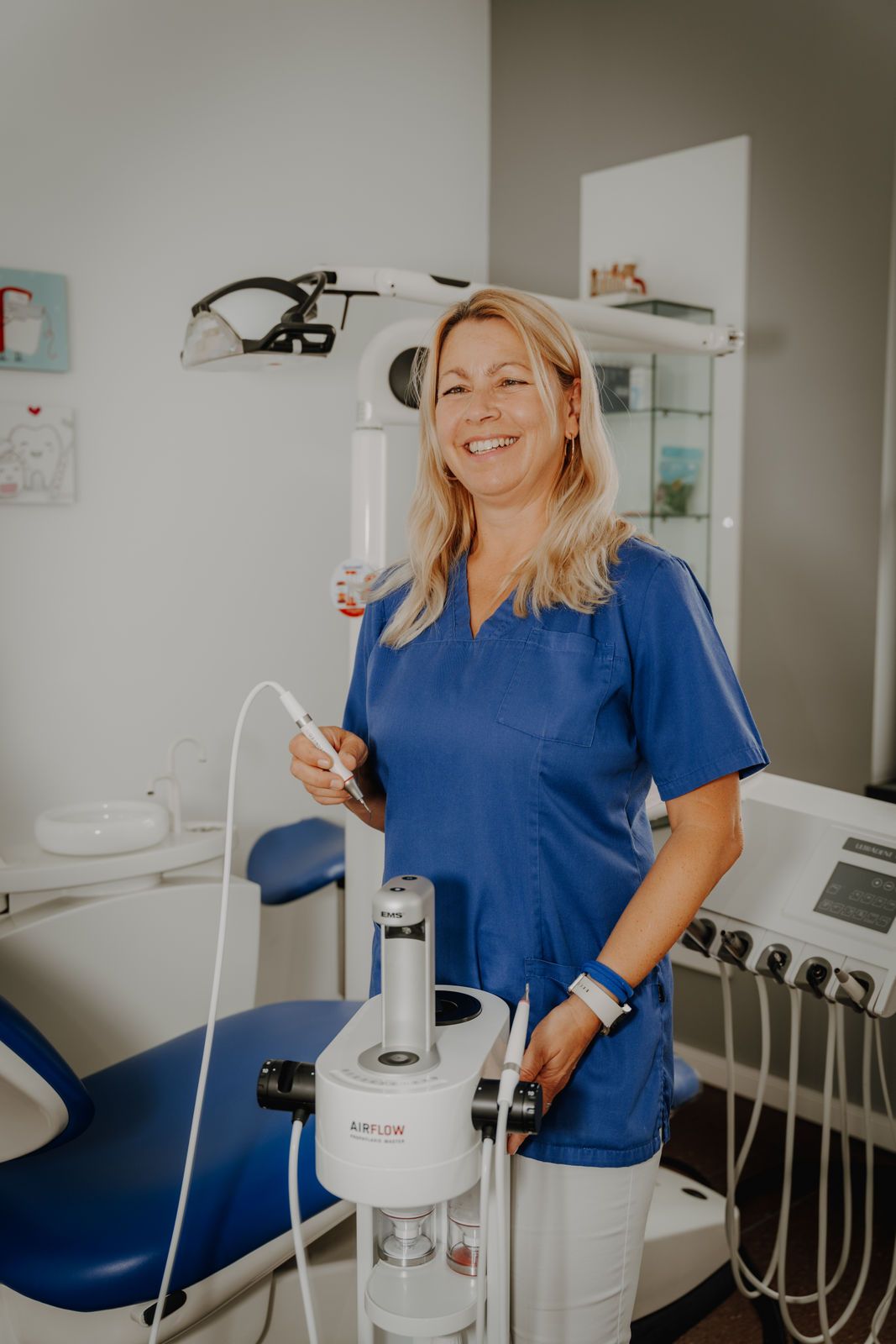 Eine Frau in blauer OP-Kleidung lächelt in einer Zahnarztpraxis und hält ein medizinisches Instrument in der Nähe von Geräten.