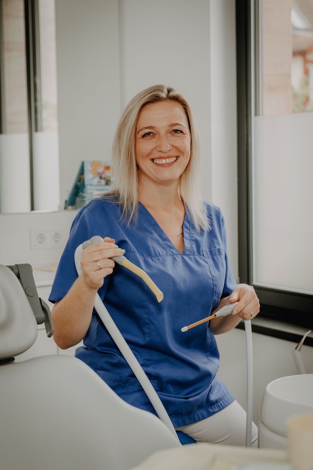 Eine lächelnde Frau in blauer OP-Kleidung hält zahnärztliche Instrumente in der Hand und sitzt auf einem Zahnarztstuhl in einer Klinik.