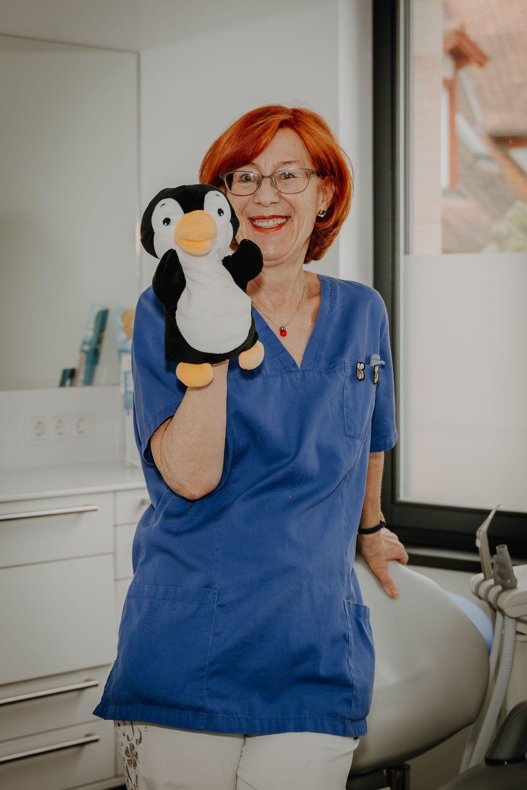 Eine Frau in blauer OP-Kleidung lächelt und hält in einer Zahnarztpraxis ein Pinguin-Stofftier in der Hand.