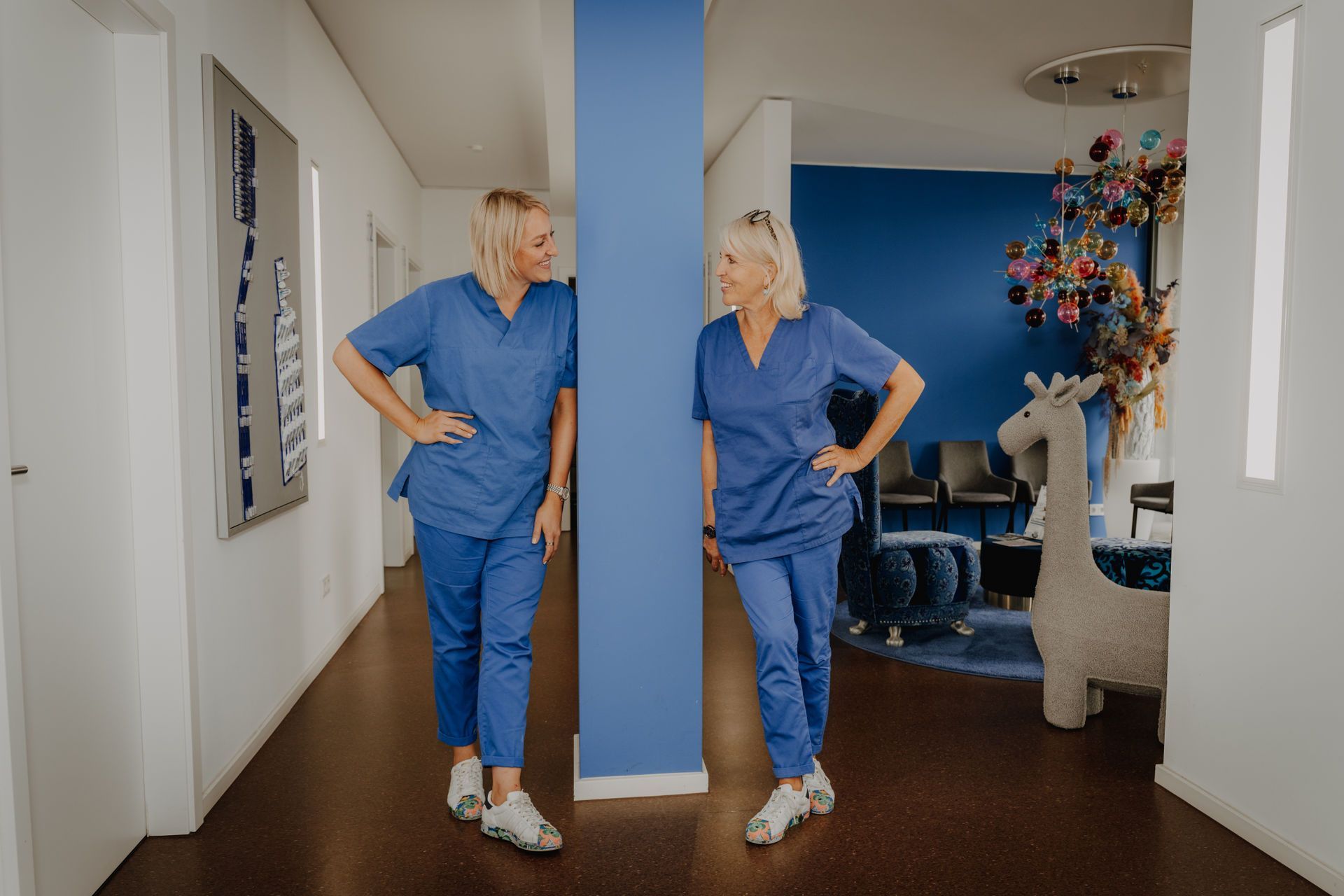 Zwei Personen in blauer OP-Kleidung lehnen lächelnd an einer blauen Säule im Flur einer Arztpraxis.