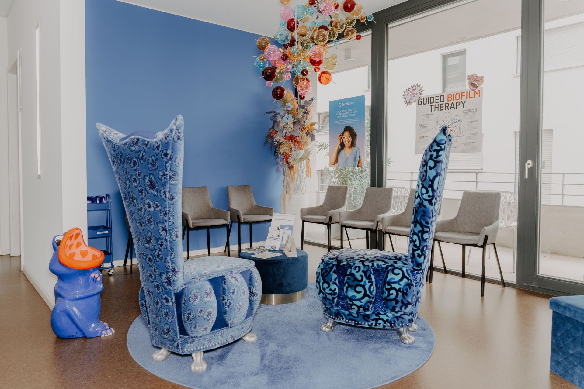 Zahnärztlicher Untersuchungsraum mit Stuhl, blauem Hocker, kleinem blauen Stuhl und Fenstern.