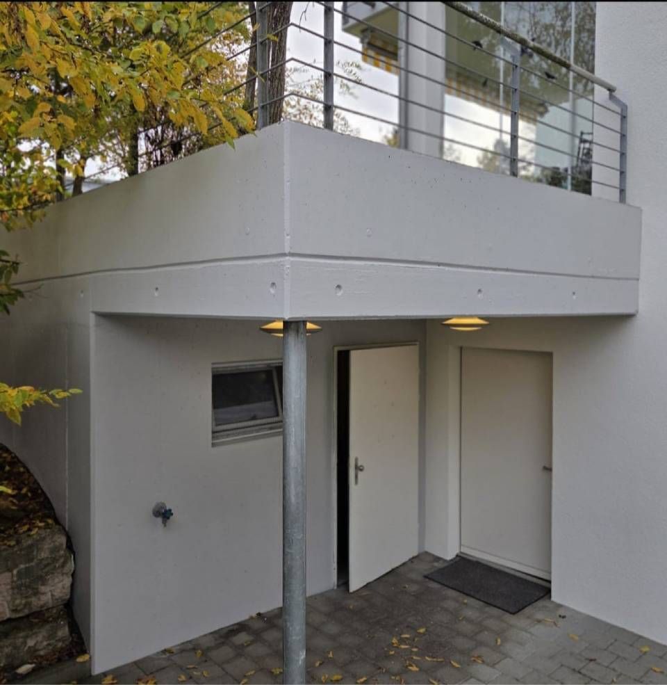 Moderner Hauseingang mit weißen Wänden, offener Tür und darüberliegendem Balkon, von einer kleinen Terrasse aus betrachtet.
