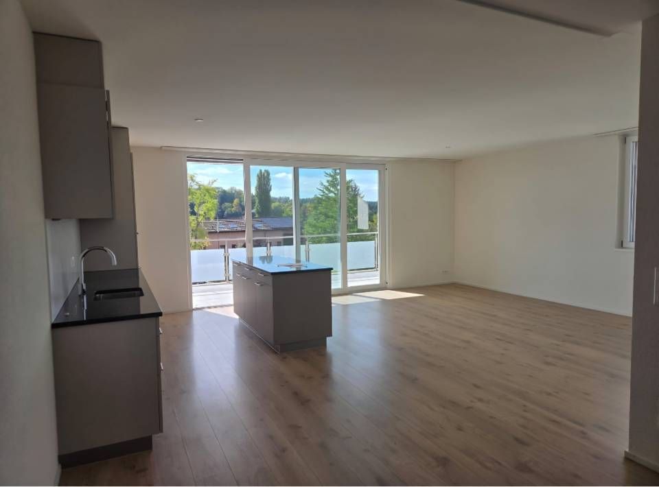 Leeres, modernes Wohnzimmer mit Holzboden, Kücheninsel und großen Glastüren zu einer Terrasse.