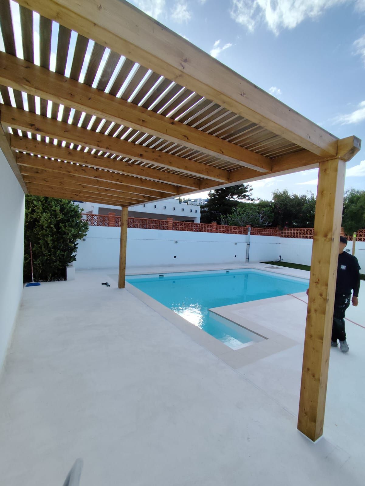 Un hombre se encuentra bajo una pérgola de madera sobre una piscina.