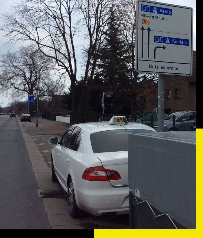 Un taxi blanco estacionado en una calle de la ciudad junto a una acera y señales de tráfico, con árboles desnudos y edificios al fondo.