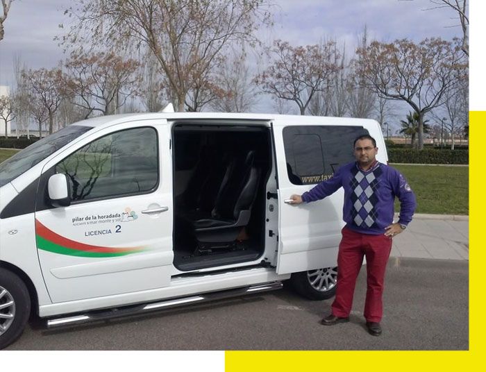 Furgoneta blanca con la puerta corredera abierta; un hombre con jersey azul y pantalones rojos está de pie junto a ella en el exterior.