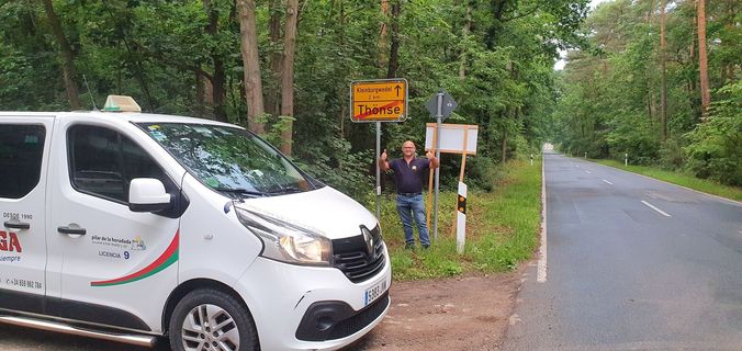A person stands by a yellow German town sign on a forested road next to a white van.